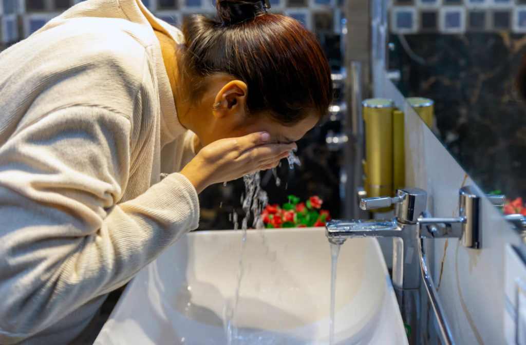 A woman in a gray colored sweatshirt is in front of a  vanity sink washing her face and eyes in running water from the faucet to remove the dirt built and to avoid MGD.