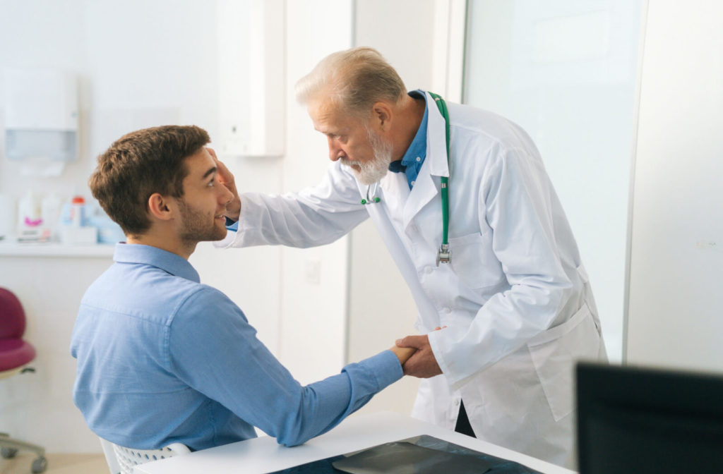 A male senior doctor in a white laboratory gown is examining the eye of a male patient in a blue long-sleeve shirt. The doctor is touching the patient's eyelid on his right index finger to see further the dry eyes in preparation for a Lipiflow procedure.