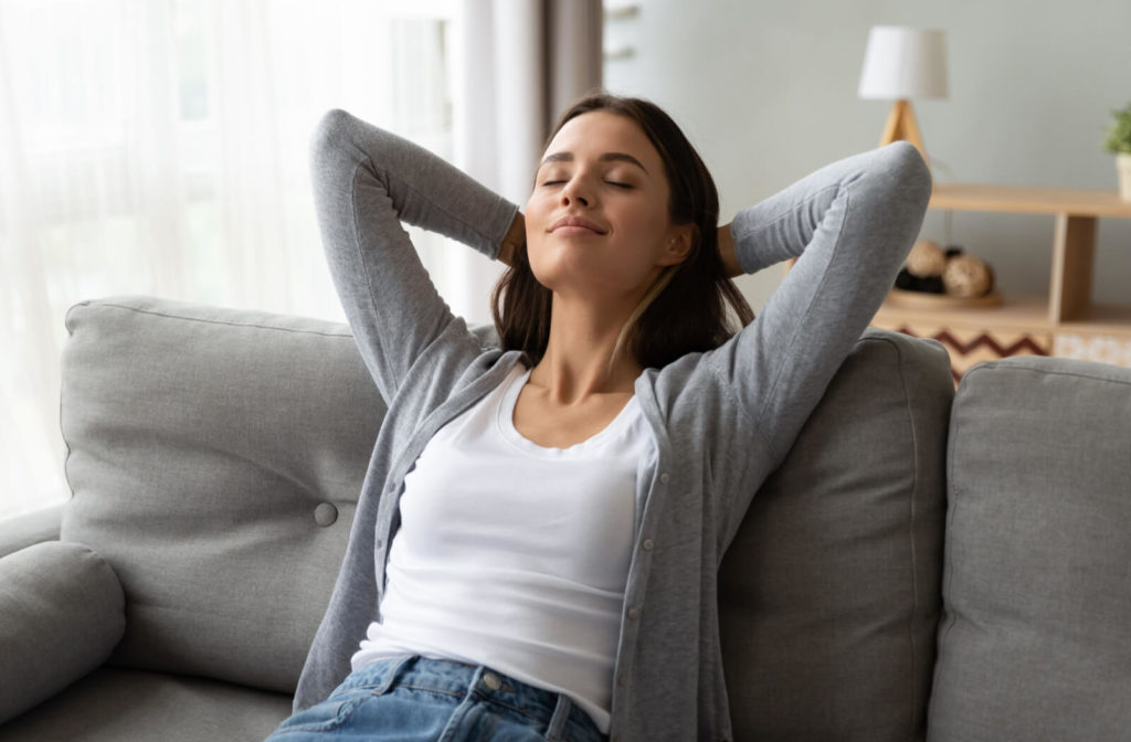 A lady in a white shirt and a gray long-sleeve cardigan with her eyes closed and feeling relaxed after her Lipiflow treatment is sitting on a gray sofa and her back leaning on the backrest.