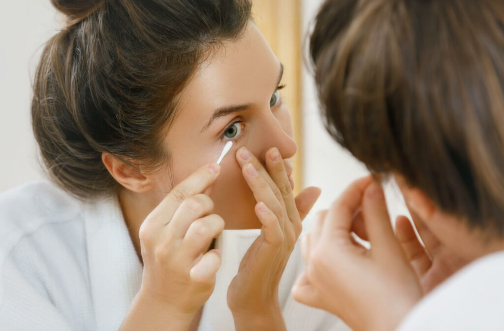 A woman in front of a mirror is cleaning her eyelid with a cotton swab.