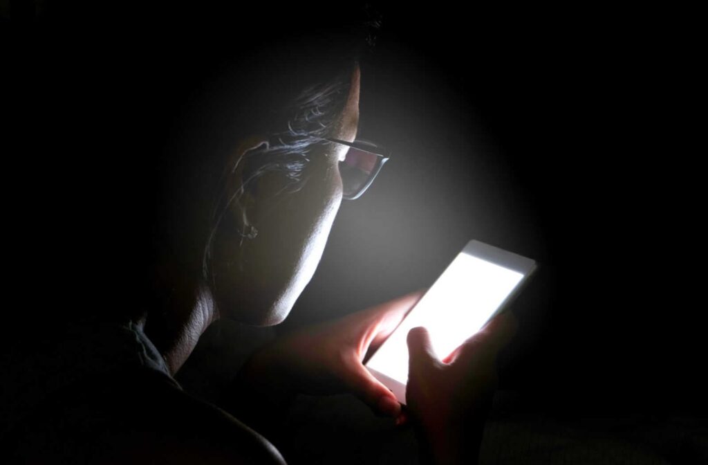 A woman is staying up late using her smartphone in her room without lights. Excessive use of digital devices like computers, tablets, and smartphones can contribute to myopia progression.