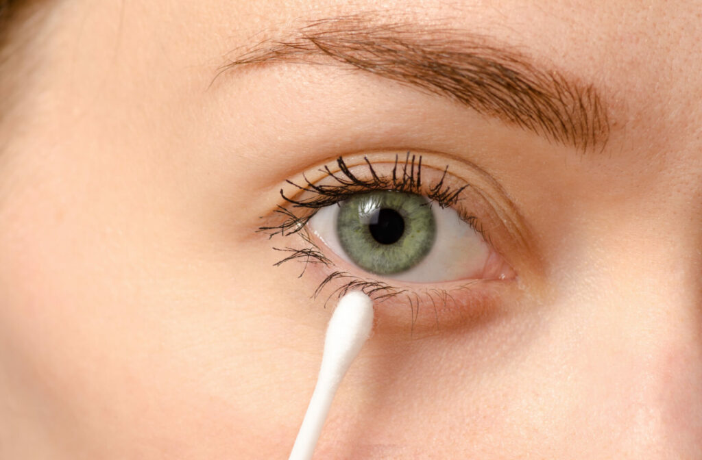 A close-up eye of a woman cleaning her eyelid with a cotton swab to remove excess mascara.