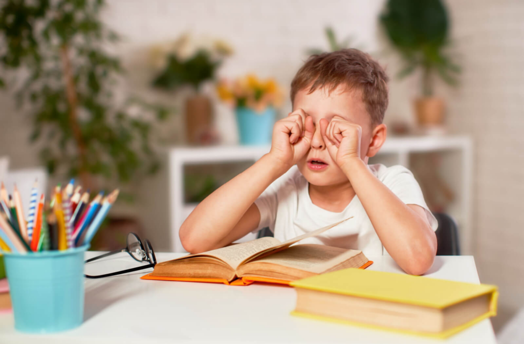 A young boy rubbing his eyes with both hands after reading the book in front of him.
