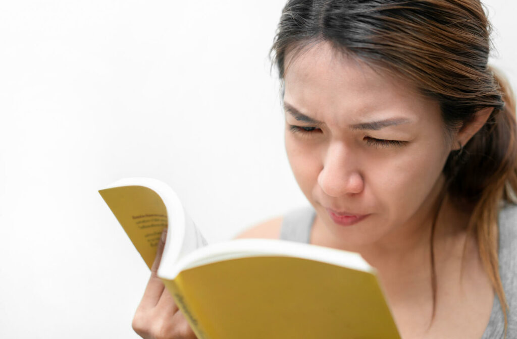 A young woman squinting her eyes to see more clearly while holding a book in her right hand.
