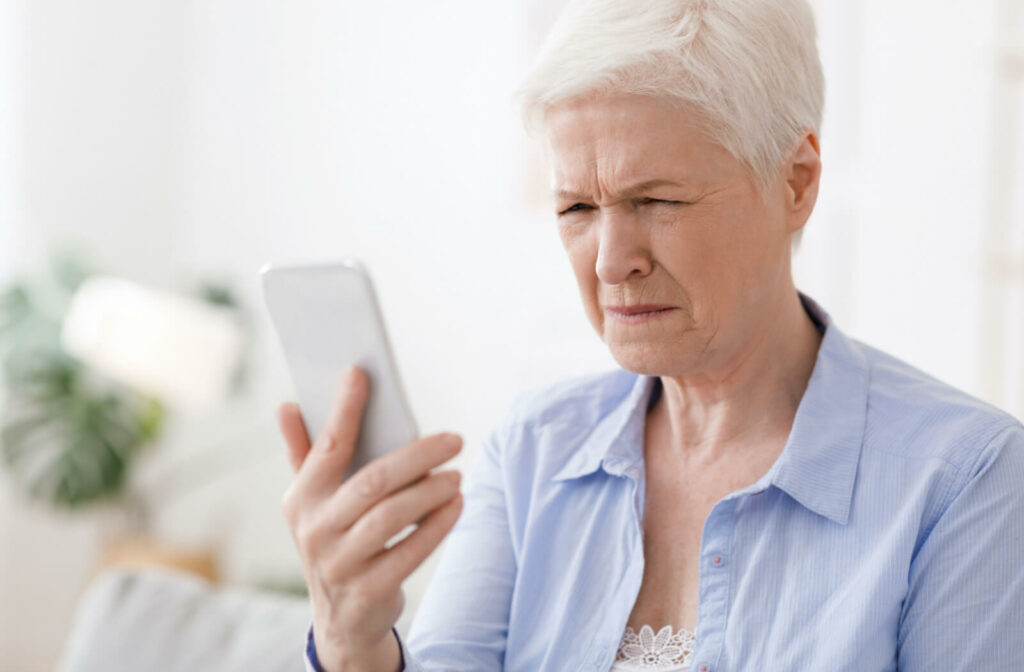 A senior woman squinting her eyes to see more clearly while holding a phone in her right hand.