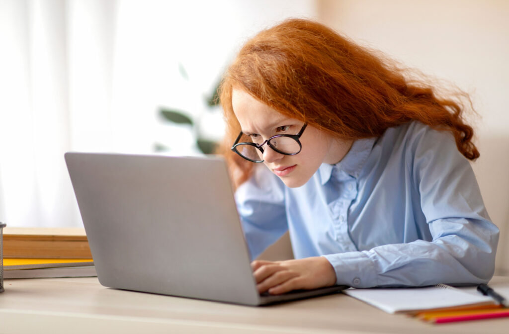 A red-haired young girl with glasses leaning closer to her laptop screen to see its content better.