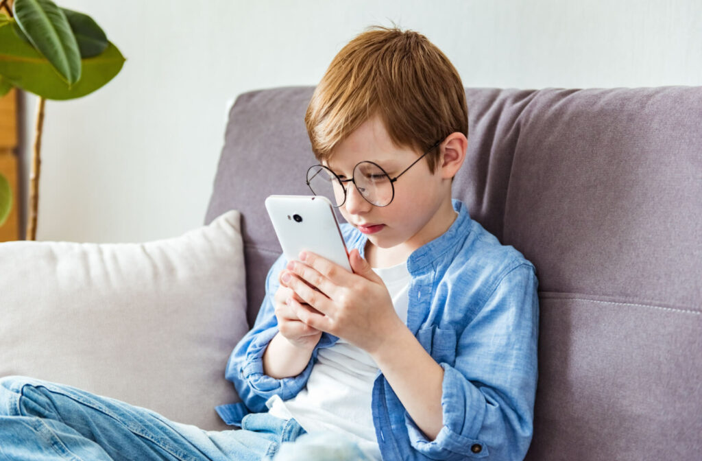 A child with large round glasses sitting on a couch and holding a smartphone very close to his face.