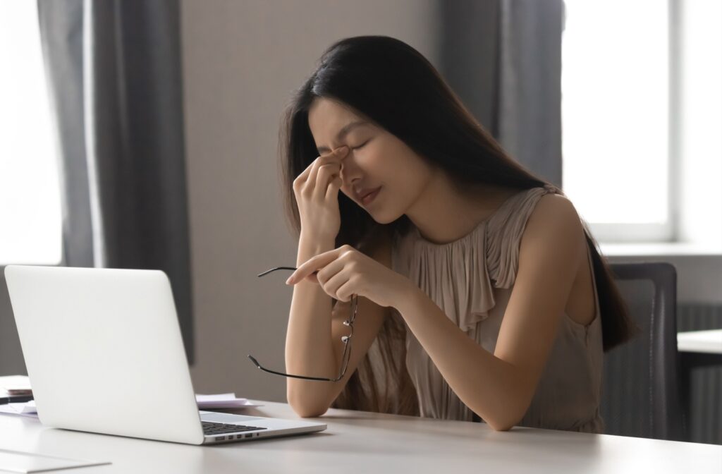 A woman sits at a desk in front of a laptop and touches her forehead with her left hand as she experiences a headache.