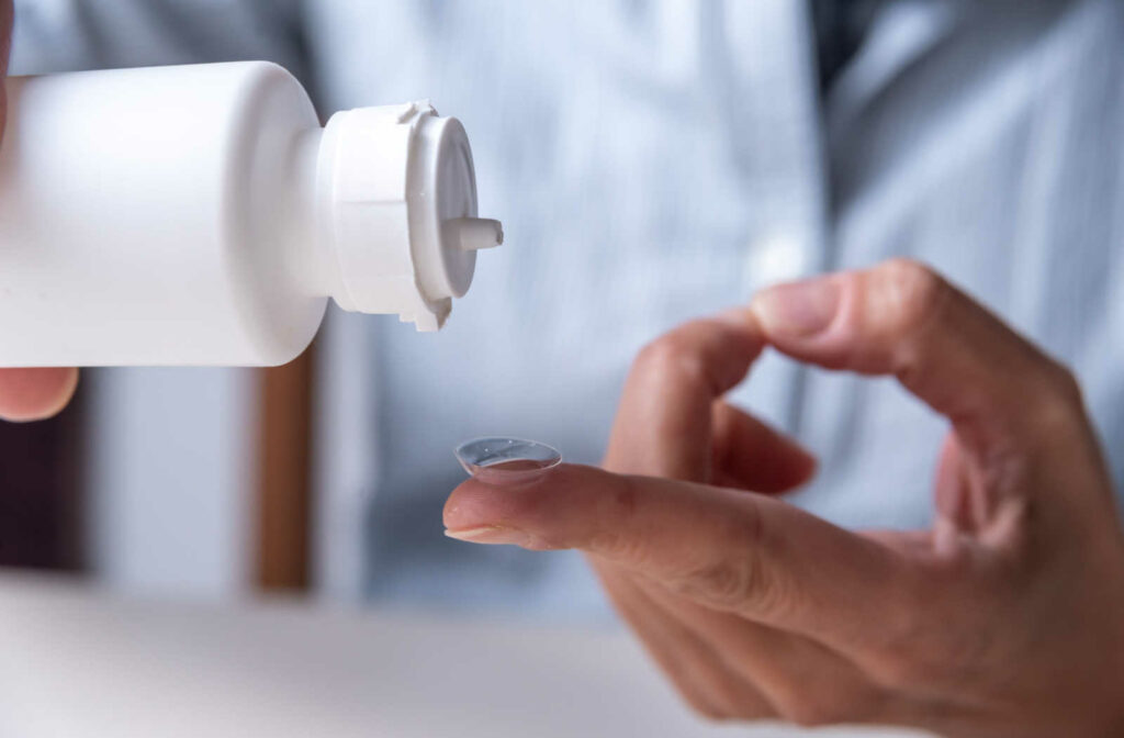 A contact lens on a fingertip with a hand pouring contact solution onto it.