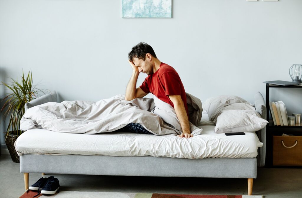 Man waking up with dry eyes and blurry vision, sitting on the edge of his bed and rubbing his forehead in discomfort.